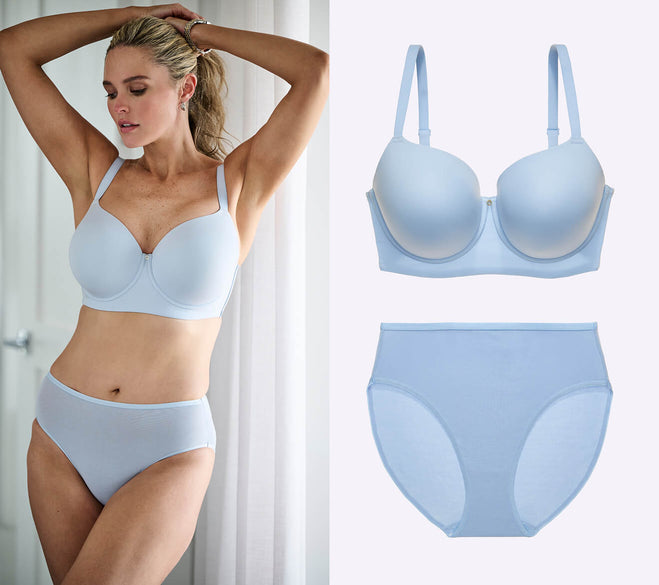 A model poses in light blue lingerie, showcasing a bra and high-waisted panties. The background features soft lighting and curtains.