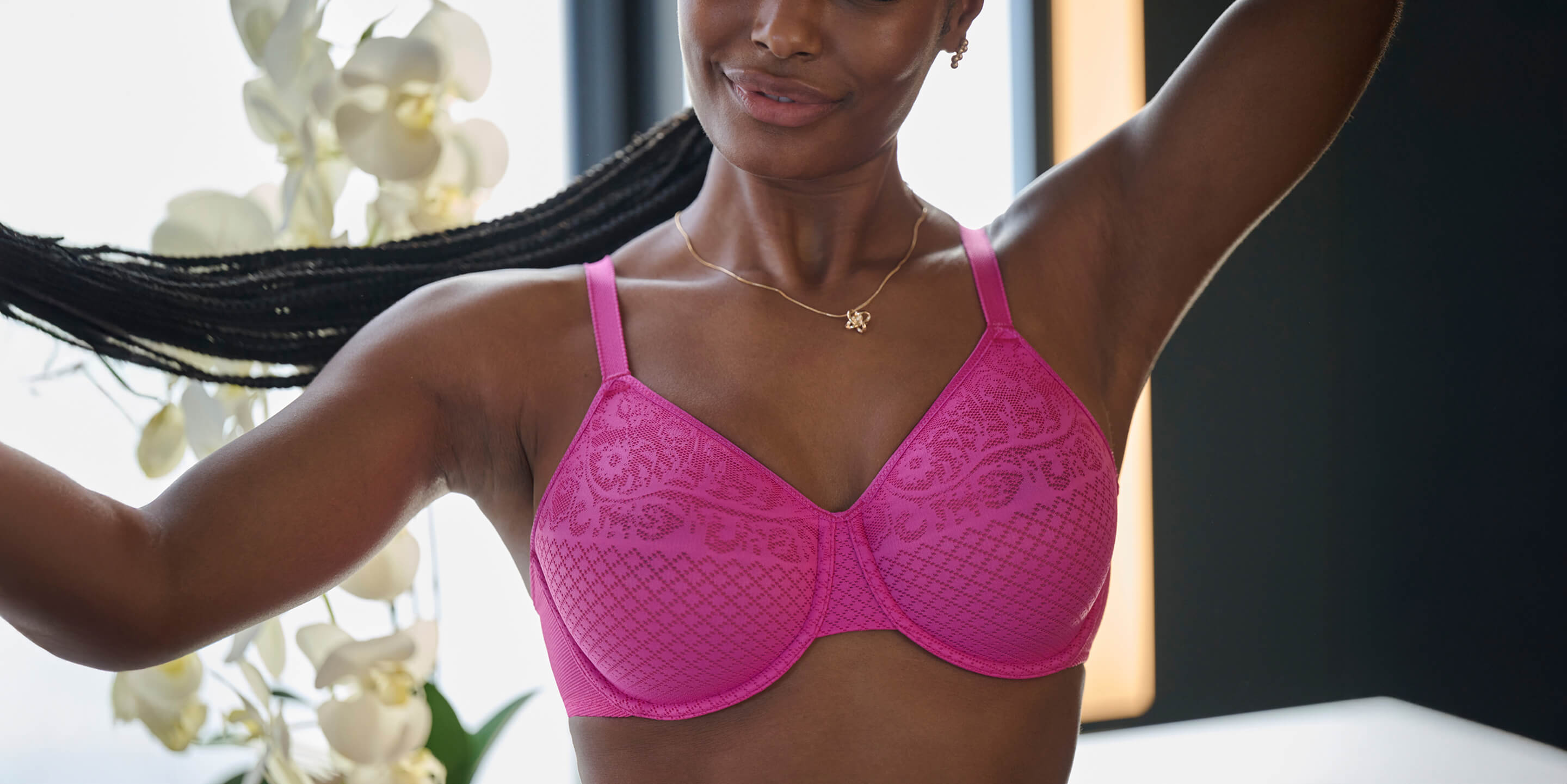 A woman poses with her arms raised, wearing a bright pink bra, with a backdrop of white orchids and soft lighting.