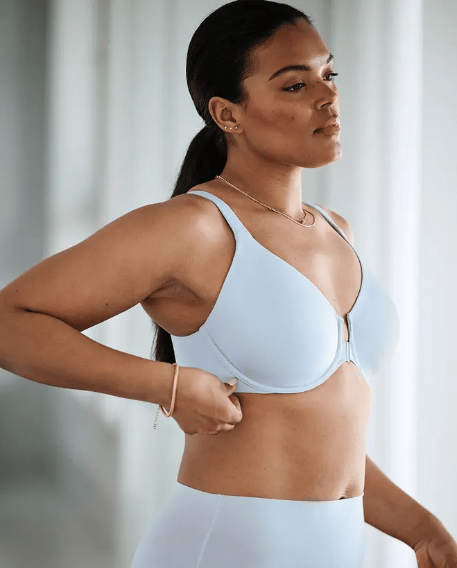 A woman adjusts her light blue bra, looking thoughtfully to the side, with a soft background.