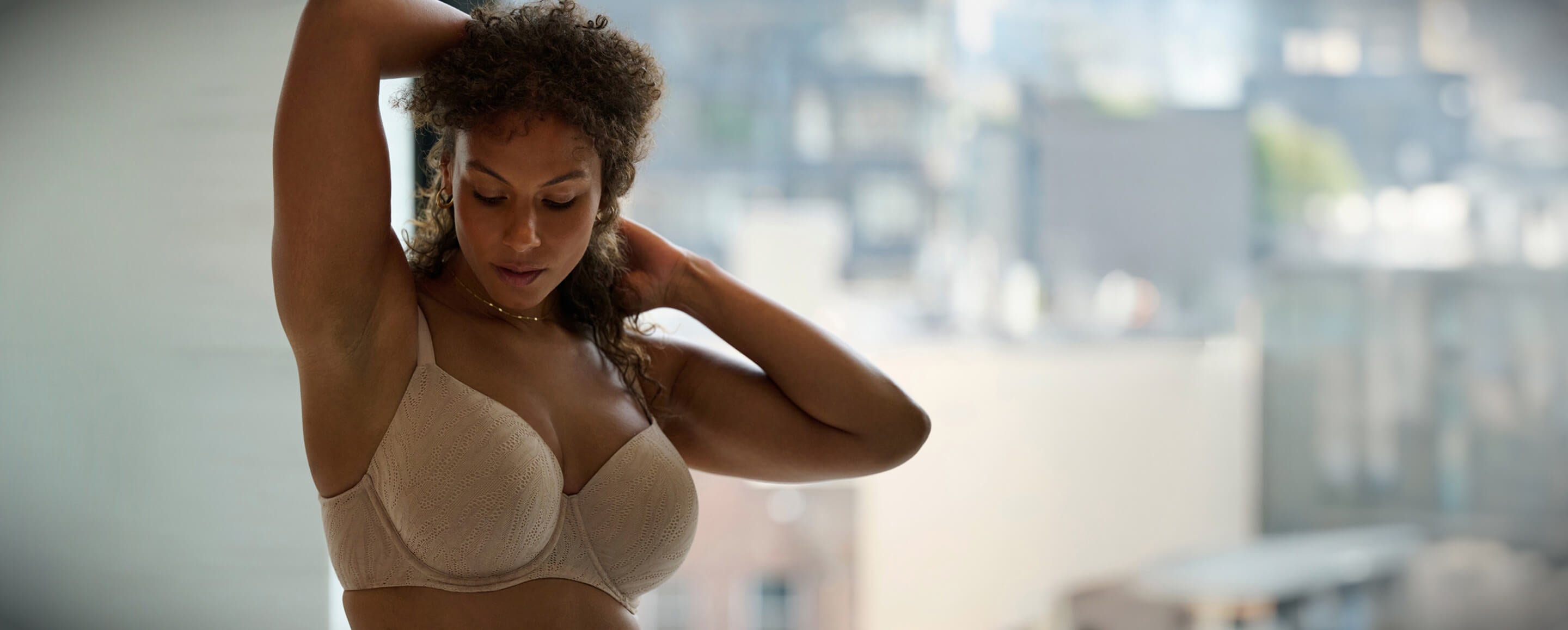 A woman with curly hair adjusts her hair while wearing a beige bra, standing in a softly lit room with a city view.